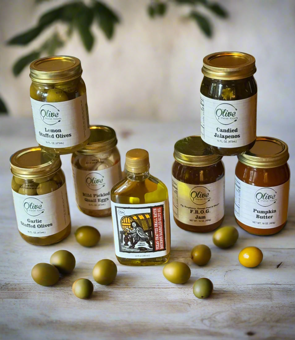 Olive These Foods small-batch jars of olives, jams, pickles, and olive oil displayed on a wooden tray with greenery.
